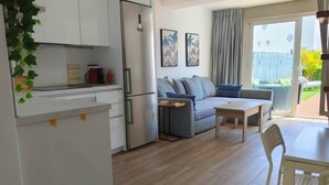 Un espacio habitable compacto con kitchenette, un sofá gris, una mesita y un ventanal corredero que da a un área exterior.