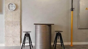 Een kamer met een metalen tonvormige tafel, twee zwarte krukken, een klok aan de muur en een gele paal.