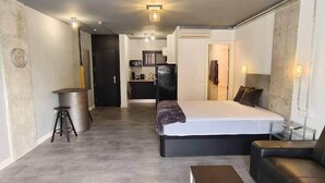 Een moderne slaapkamer met een bed, een kleine bank, een bijzettafeltje en een kitchenette op de achtergrond.