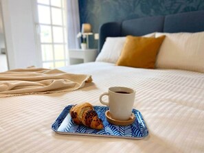 Ein ordentlich bezogenes Bett mit cremefarbener Decke, ein blau-weiß gemustertes Tablett mit einem Croissant und einer Kaffeetasse sowie eine beigefarbene Tagesdecke.