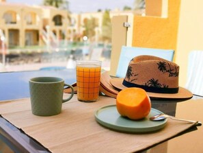Ein Frühstückstisch mit Tasse, Orangensaftglas, einem Teller mit Obstscheibe und einem Hut, mit Blick auf einen Pool.
