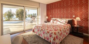 A bedroom with a bed, two bedside tables, a nightstand, a chair, and a view through a sliding glass door to a balcony.