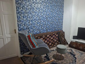 Une pièce avec un mur bleu à motifs, un fauteuil gris agrémenté d’un plaid coloré, un canapé à motifs, un petit pouf et un téléviseur posé sur un meuble.