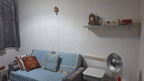Una sala de estar con un sofá azul, una mesita, un radiador, un estante montado en la pared con adornos y un ventilador de pie.