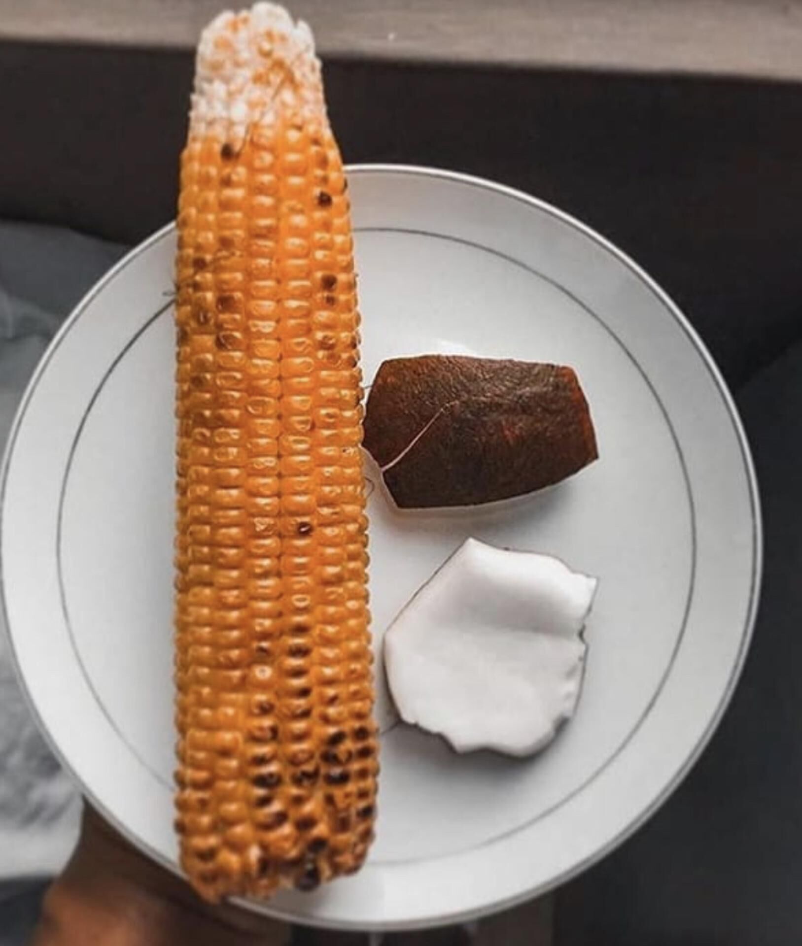 Ein Teller mit einer einzelnen Maiskolben, einem Stück dunkelbraunem Fleisch und einem Klecks weißer Sauce.
