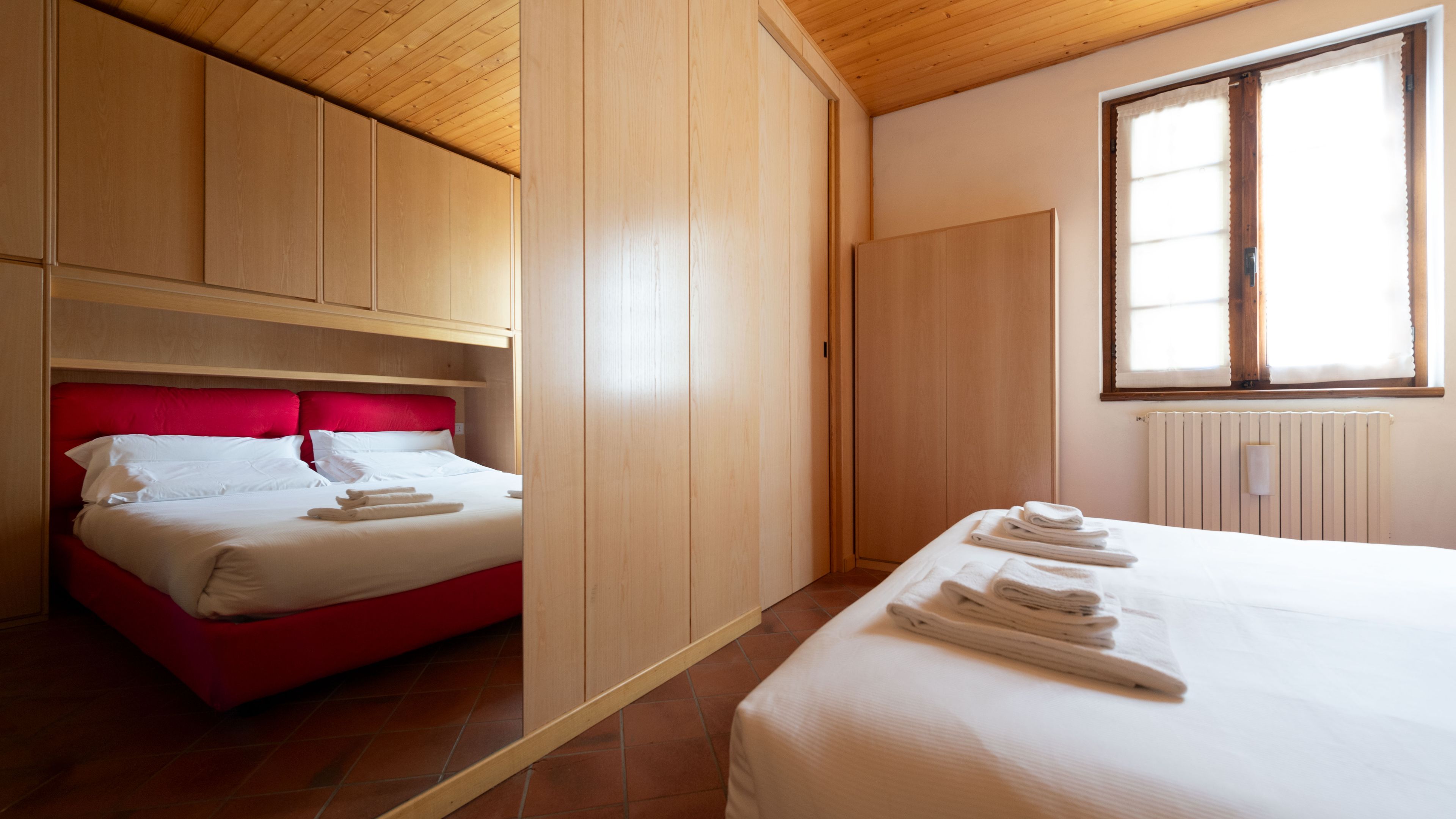 A hotel room with a bed, a red headboard, wooden paneling, and a window with curtains.