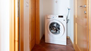 Une petite machine à laver à chargement frontal, de couleur blanche, installée dans une petite buanderie avec des portes en bois et un sol carrelé.