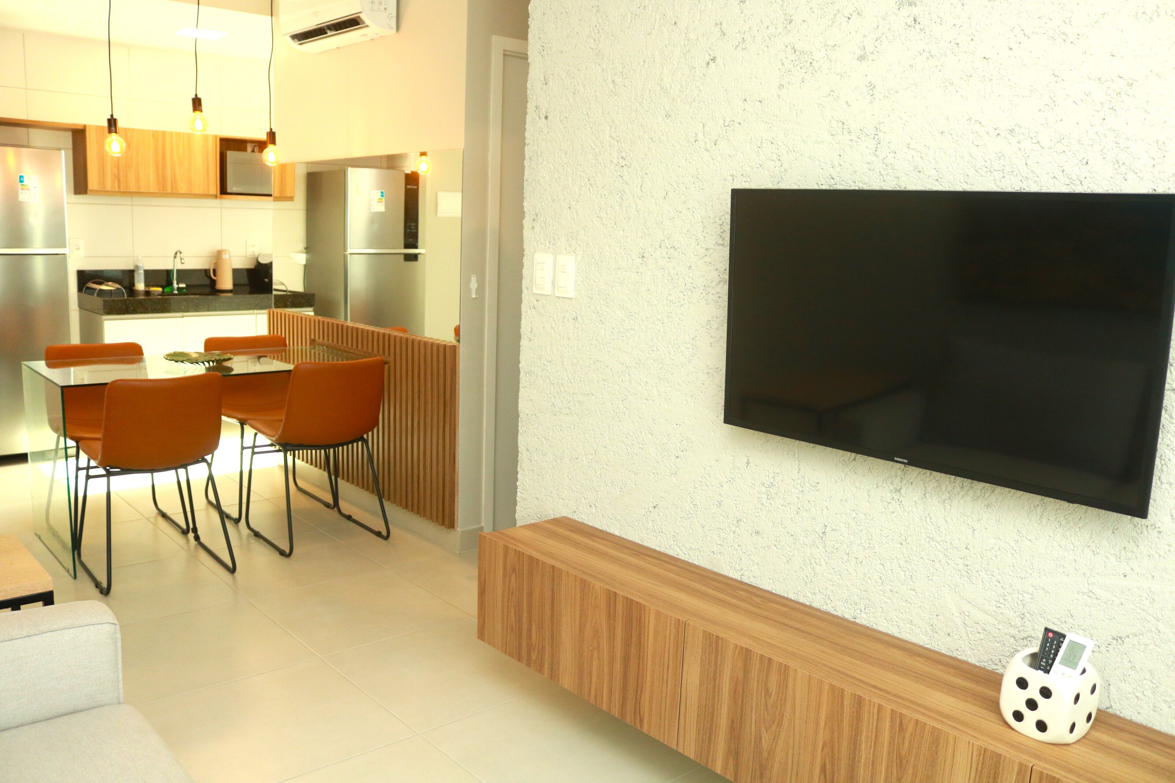 A modern kitchen with stainless steel appliances, a dining area with orange chairs, and a flat-screen TV mounted on the wall.
