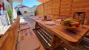 An outdoor dining area with wooden benches and tables, a basket of fruit, and a grill in the background.