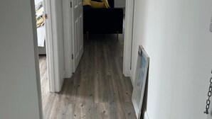 Ein schmaler Flur mit Holzboden, einem Bett mit gelben Kissen und einem kleinen Fenster mit Blick nach draußen.