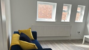 Ein Zimmer mit einem dunkelblauen Sofa, zwei Fenstern und Ziegelwänden, sowie einem Heizkörper.