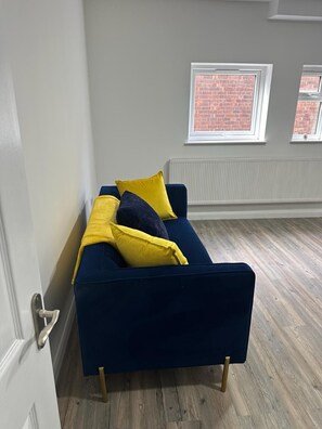 A dark blue sofa with gold legs in a room with wooden flooring, white walls, and two windows with brick views. - Immaculate 1-bed Apartment in Northampton (Northampton)