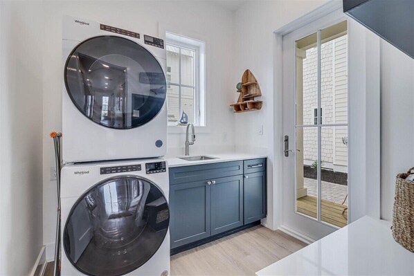 A laundry room with a front-loading washer and dryer, a sink, and a small wooden shelf.