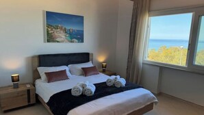 A neatly made bed with white linens and rolled towels, a painting of a coastal scene on the wall, and a view of the sea through a window. - 2 Bedroom Apartment in PALERMO (PALERMO)