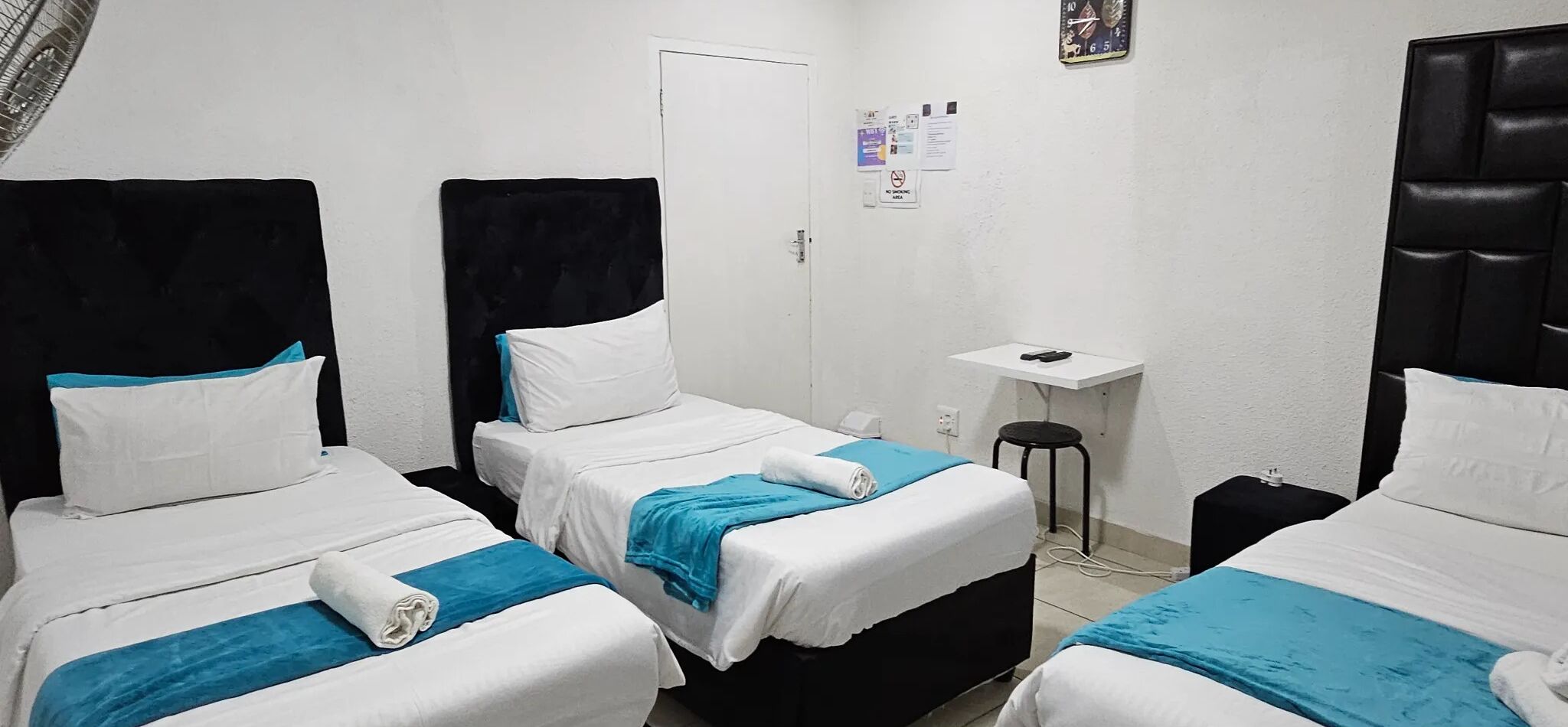 A room with three single beds, each with white bedding and a blue accent. There is a white door, a small table with a chair, and a wall-mounted clock.