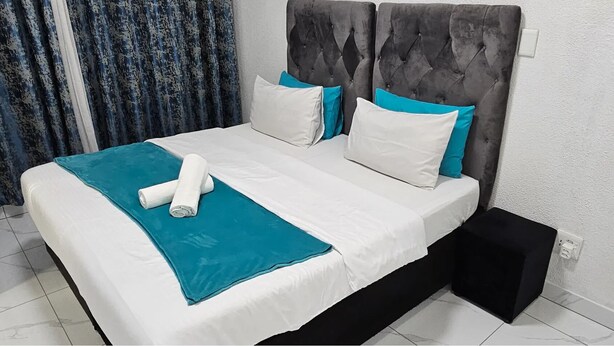 Un letto con una testiera grigia rivestita in tessuto, biancheria da letto bianca e color acquamarina, e un asciugamano bianco arrotolato.