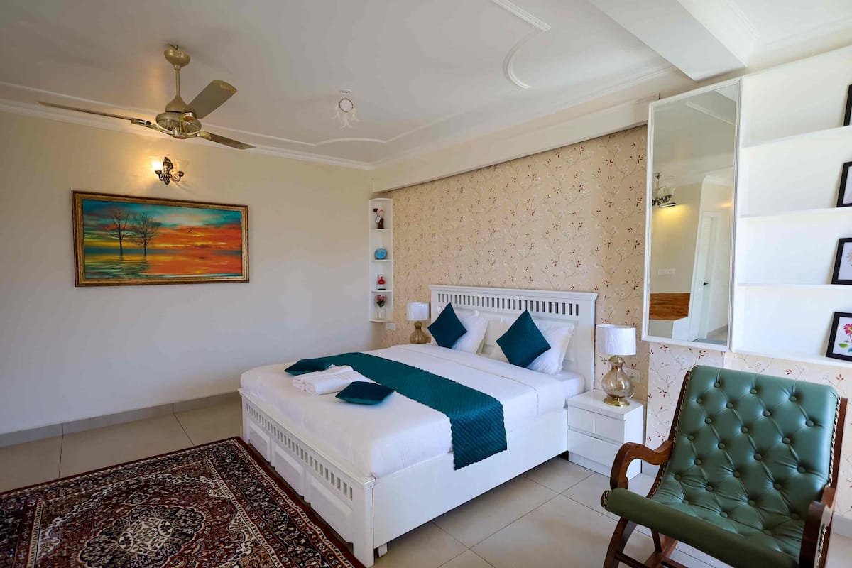 A neatly arranged bedroom with a bed, a green upholstered chair, a ceiling fan, a bedside table with a lamp, and a wall-mounted shelf with decorative items.