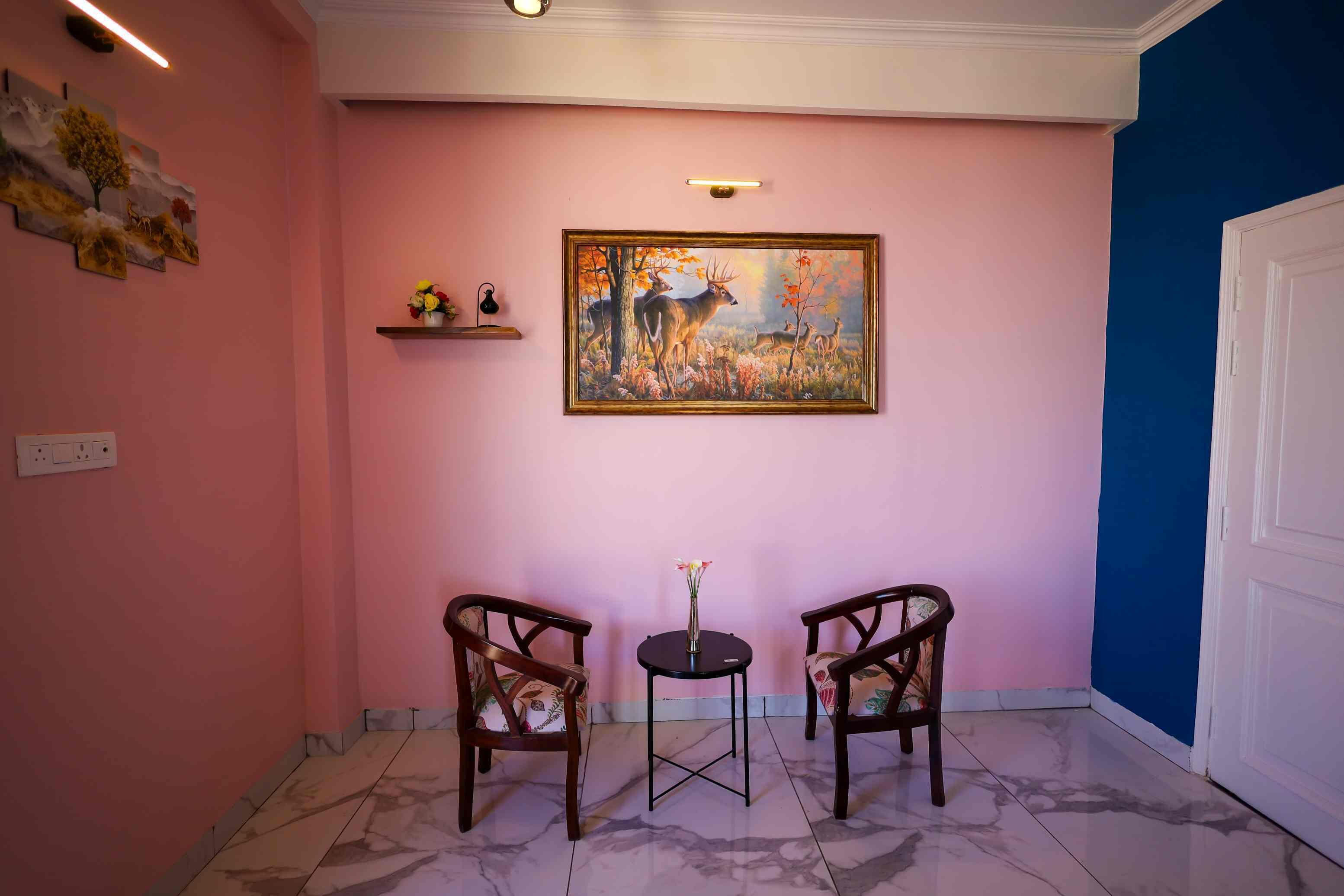 A hallway with pink walls, a painting of deer, two chairs, a small table with flowers, and a blue wall on the right.