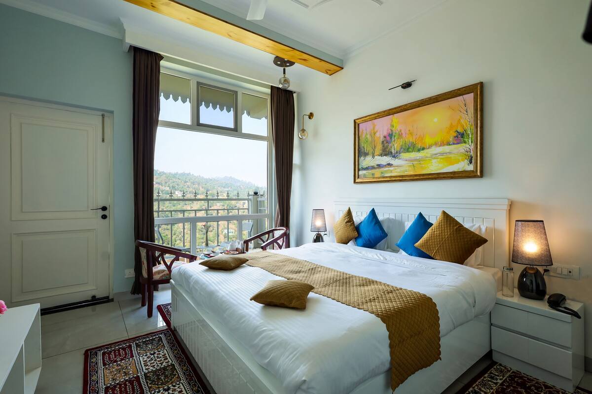 A hotel room with a large bed, a painting, a view through the window, and a balcony.