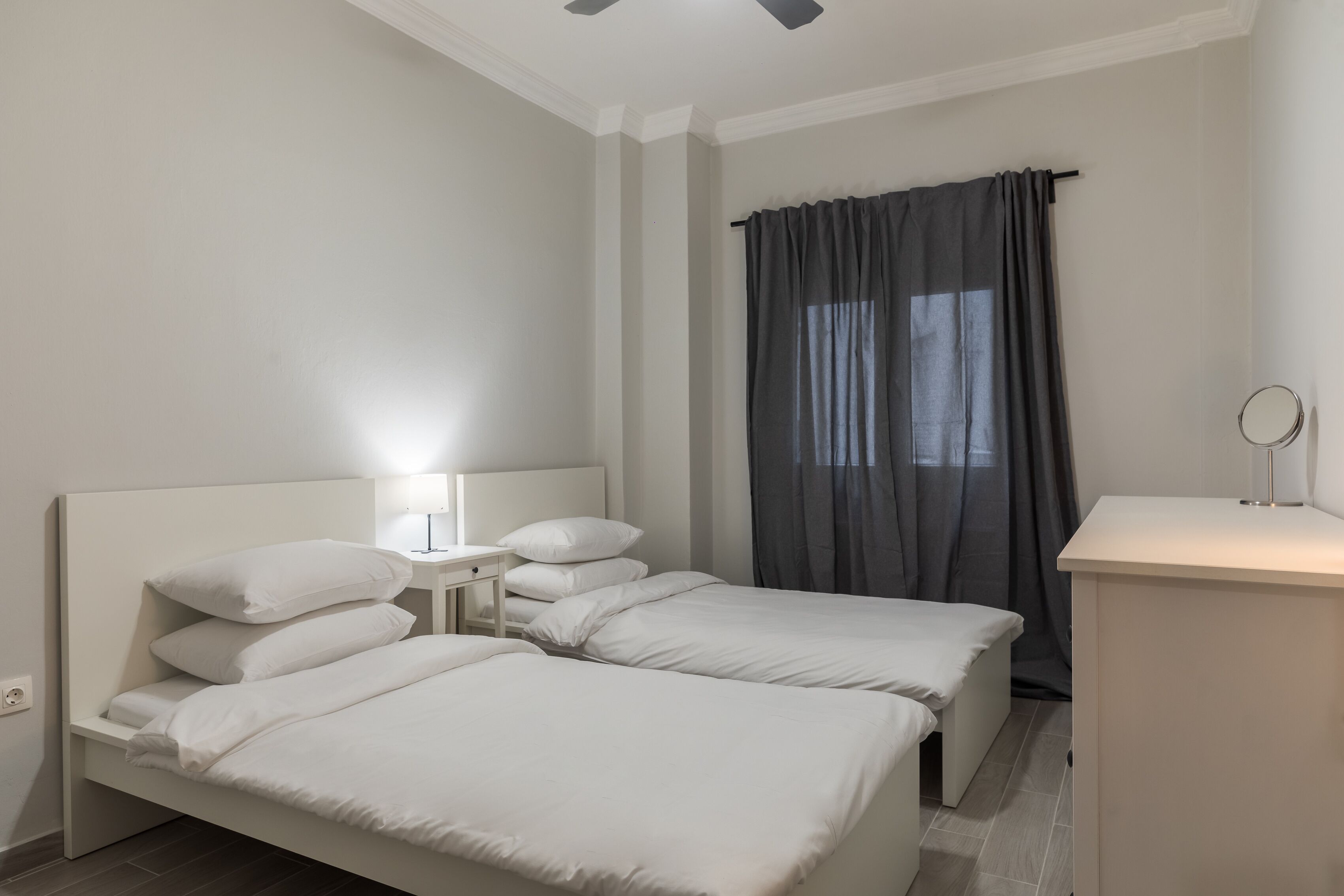 A hotel room with two single beds, a bedside table with a lamp, a small desk with a mirror, and a window with curtains.