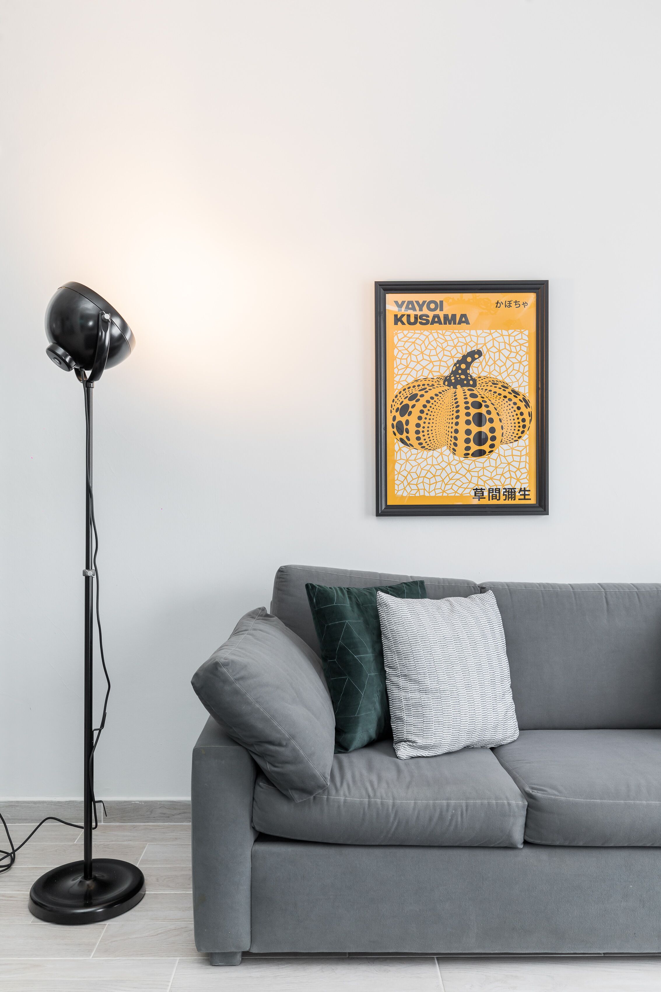 A grey sofa with two pillows, a framed artwork of a pumpkin, and a black floor lamp against a white wall.