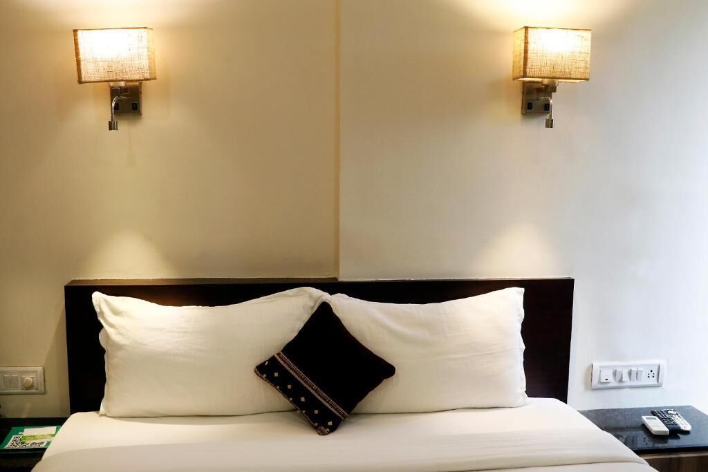 A hotel room with a neatly made bed, two wall-mounted lamps, a bedside table with a remote control, and a power outlet.