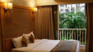 A hotel room with a wooden headboard, a large bed with white and patterned bedding, a balcony with curtains, and a view of greenery outside. - THE MYRIAD HOTEL (Mumbai)