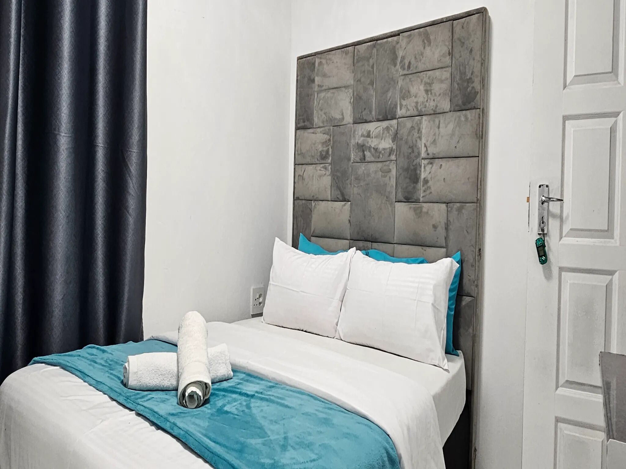 A neatly made bed with white and teal bedding, a grey headboard, and a blue curtain.