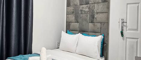 A neatly made bed with white and teal bedding, a grey headboard, and a blue curtain.