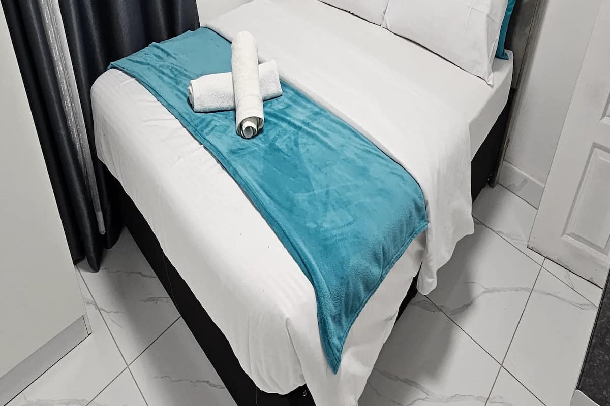 Una cama bien hecha con sábanas blancas y una manta color turquesa, una cortina negra y un piso de baldosas.