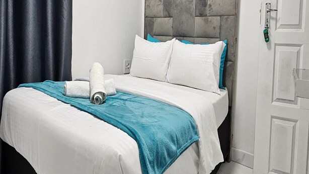 A neatly made bed with white bedding and a teal throw blanket, a white pillow, and a rolled towel on the bed. - Goshen Lodge Spencer (Cape Town)