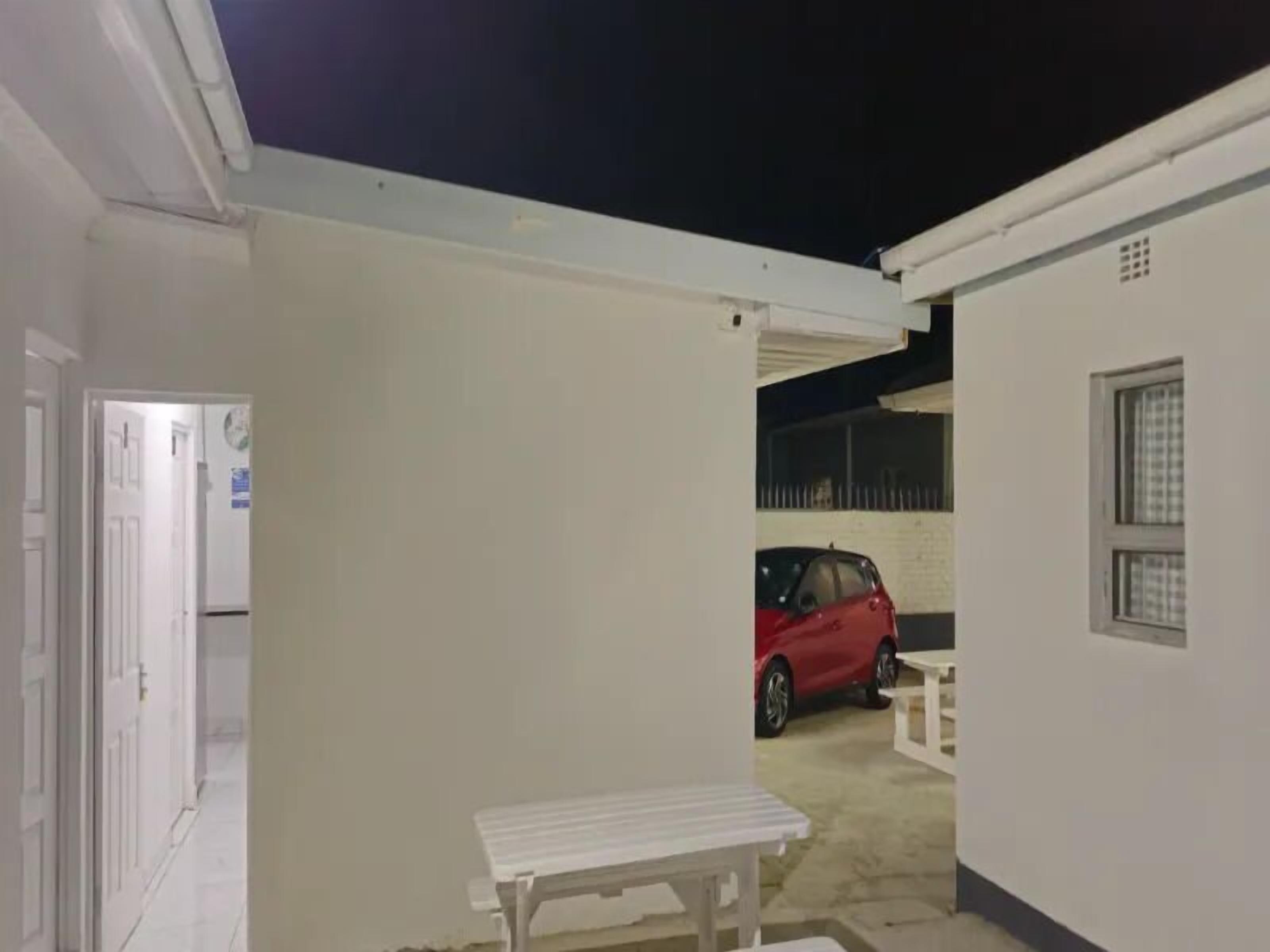 Un espacio al aire libre cubierto con un banco blanco, un coche rojo estacionado al fondo y un edificio blanco con una ventana.