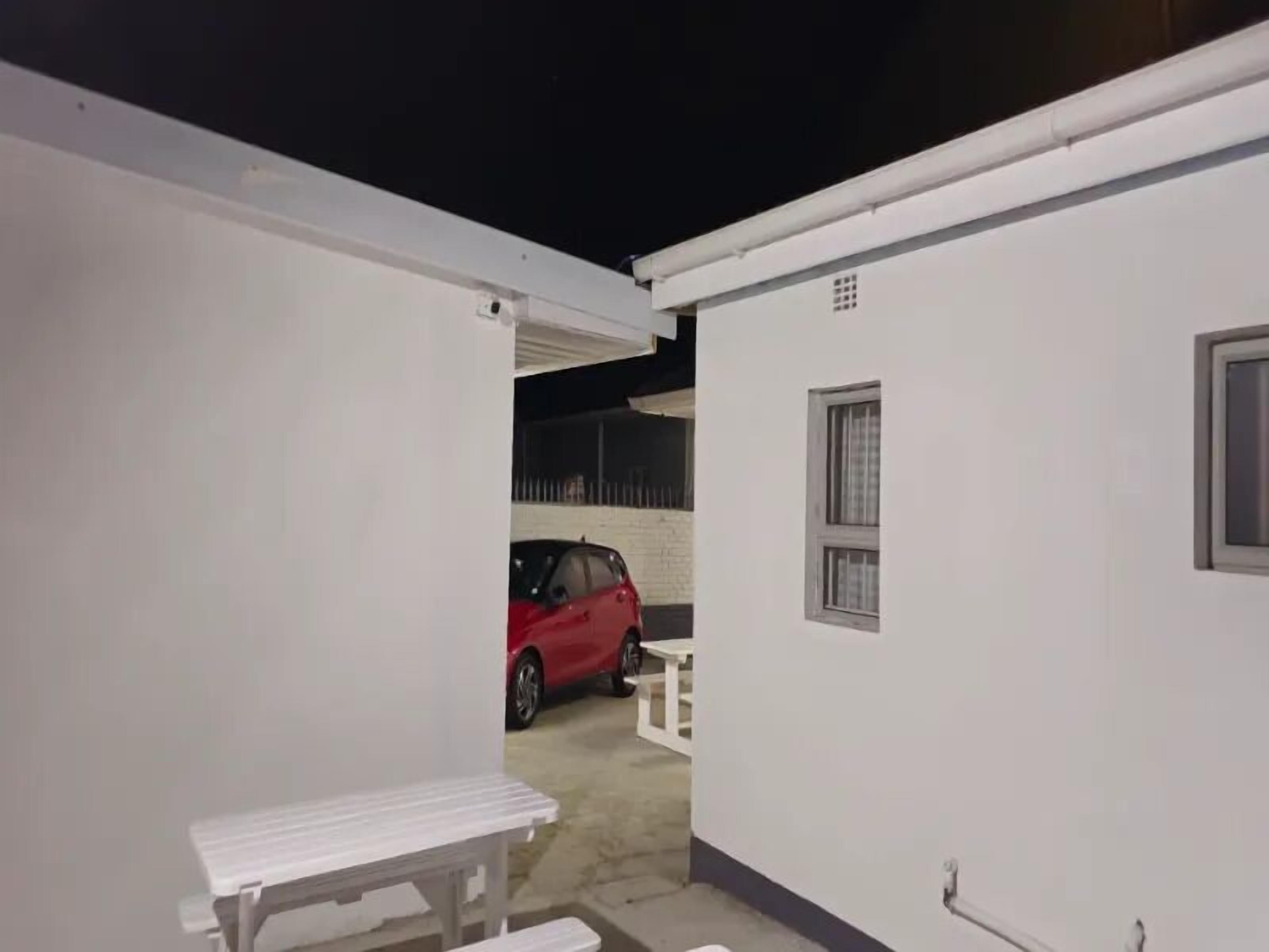 Un espacio exterior pequeño y cerrado con un banco blanco y una mesa, un coche rojo estacionado al fondo y un edificio blanco con una ventana.