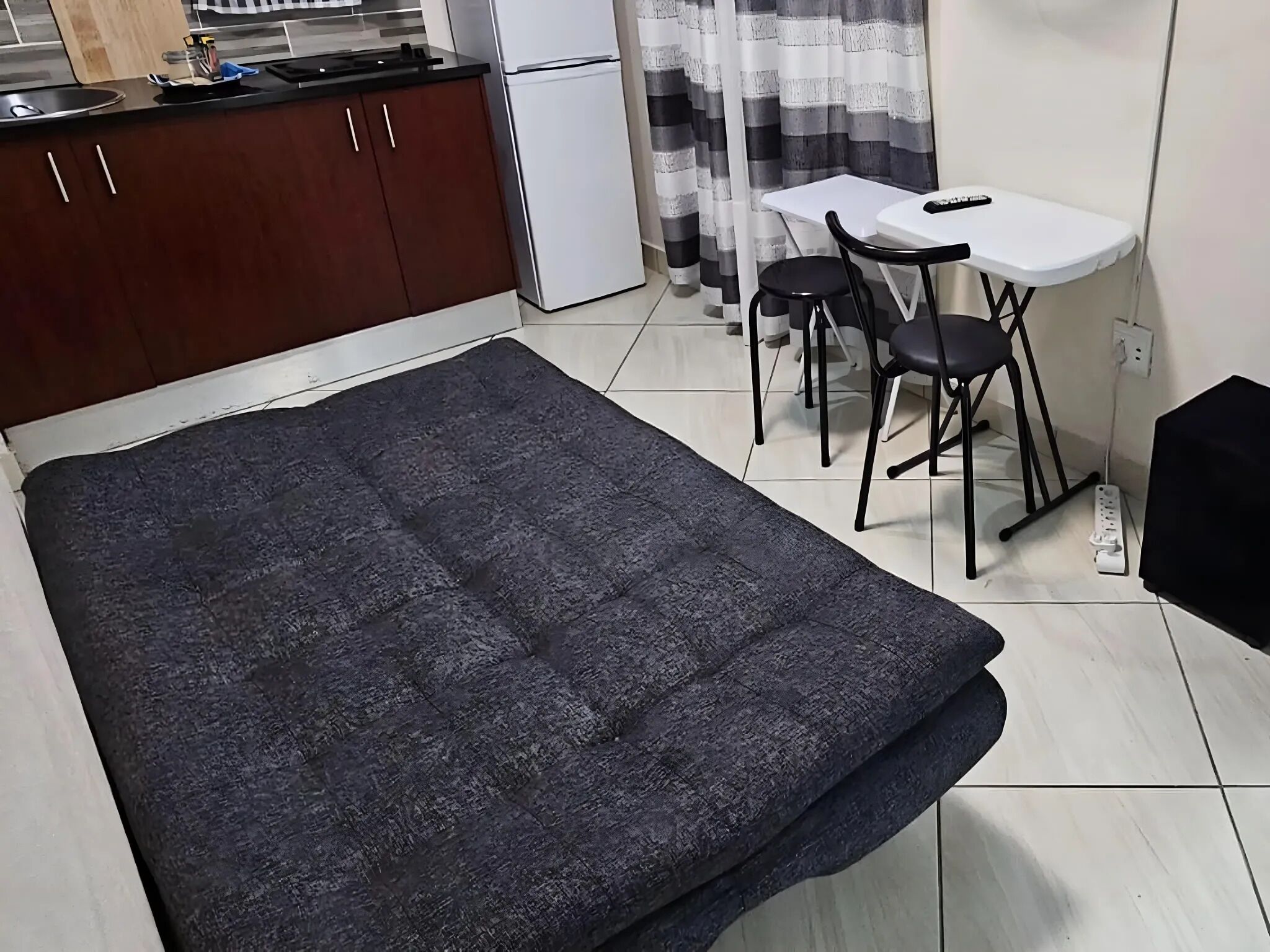 Quarto compacto com cama, pequeno kitchenette, área de refeições com mesa e cadeiras, e cortina.