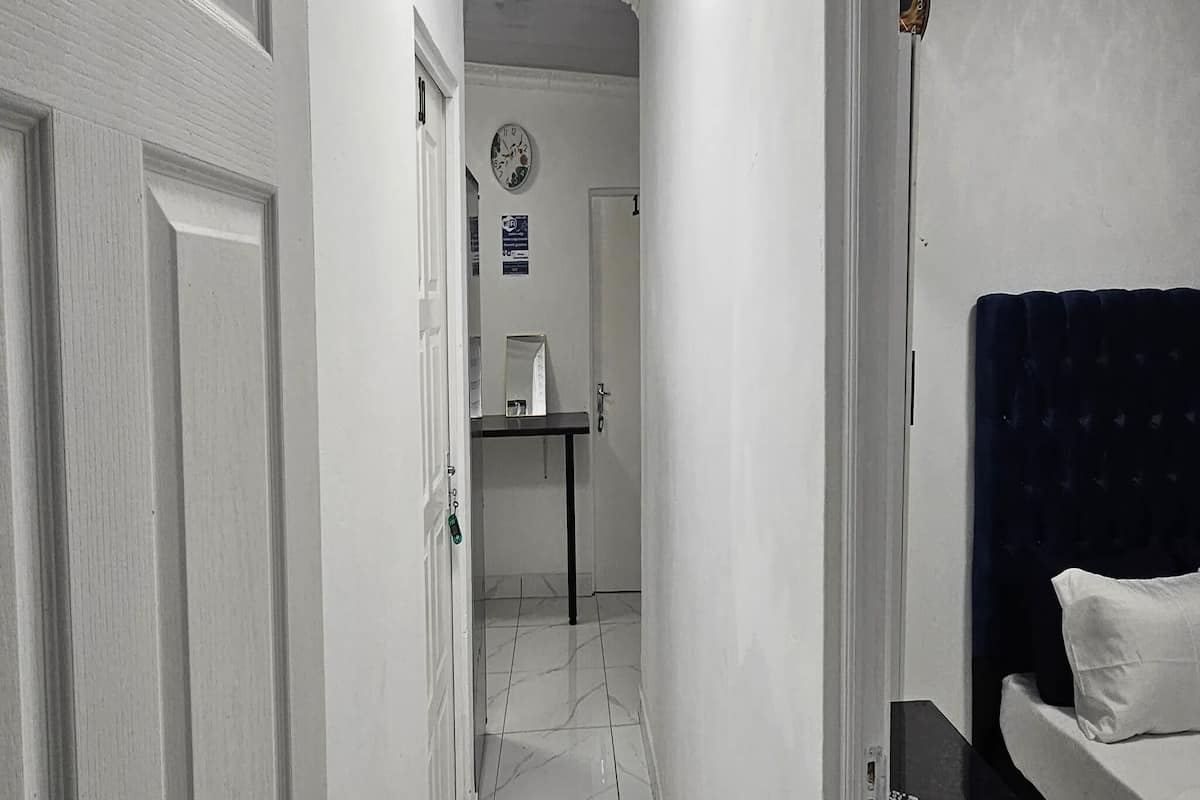 Un pasillo estrecho con una puerta a la izquierda, una pequeña mesa con un espejo y un reloj en la pared, y una cama con cabecera oscura a la derecha.