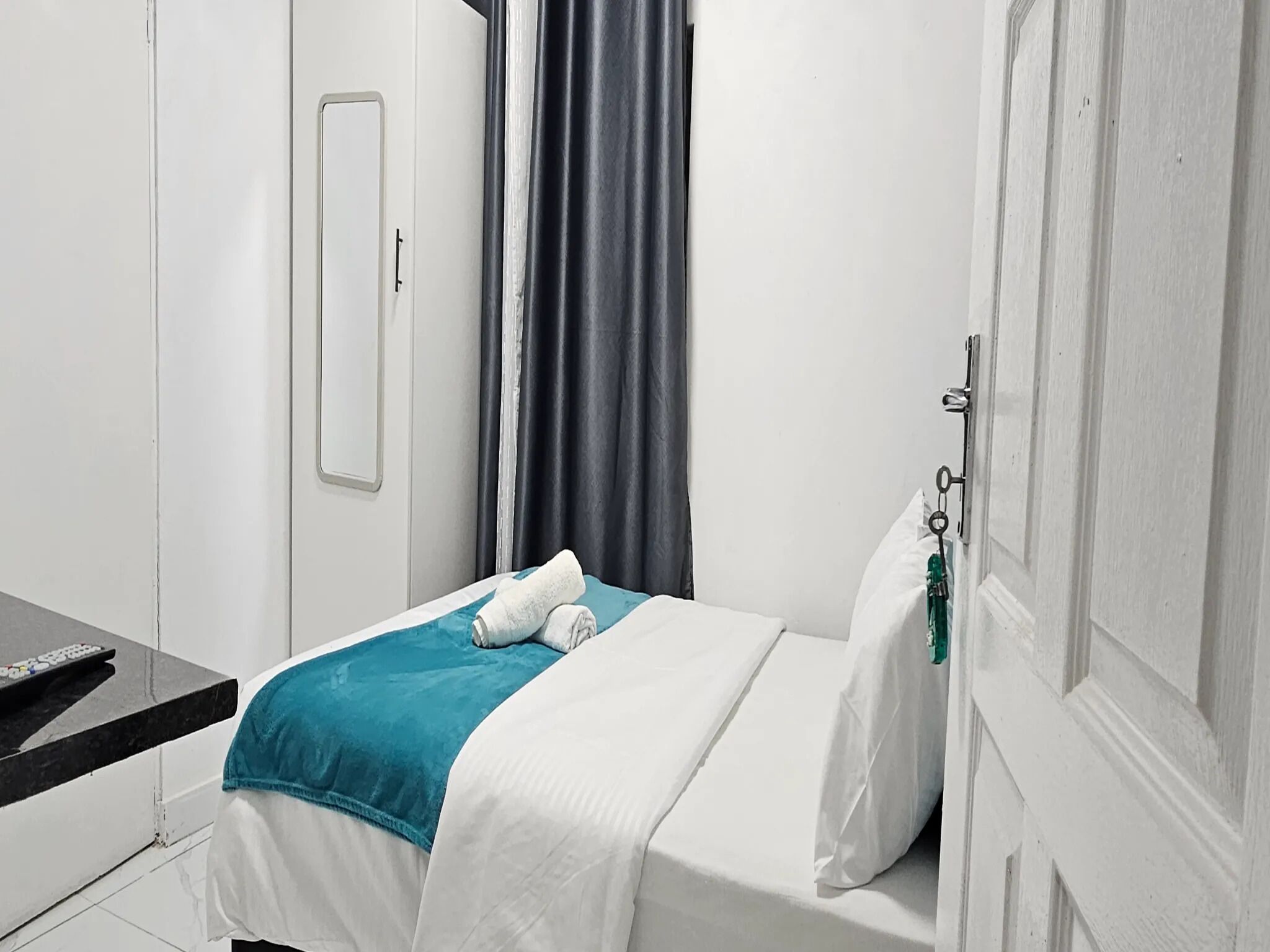A small, neatly made bed with a white and teal blanket, a single white pillow, and a remote control on a black surface beside the bed.