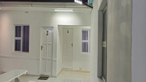 A clean, well-lit exterior of a building with three white doors, each marked with a number. There is a small outdoor seating area with benches. - Goshen Lodge Spencer (Cape Town)