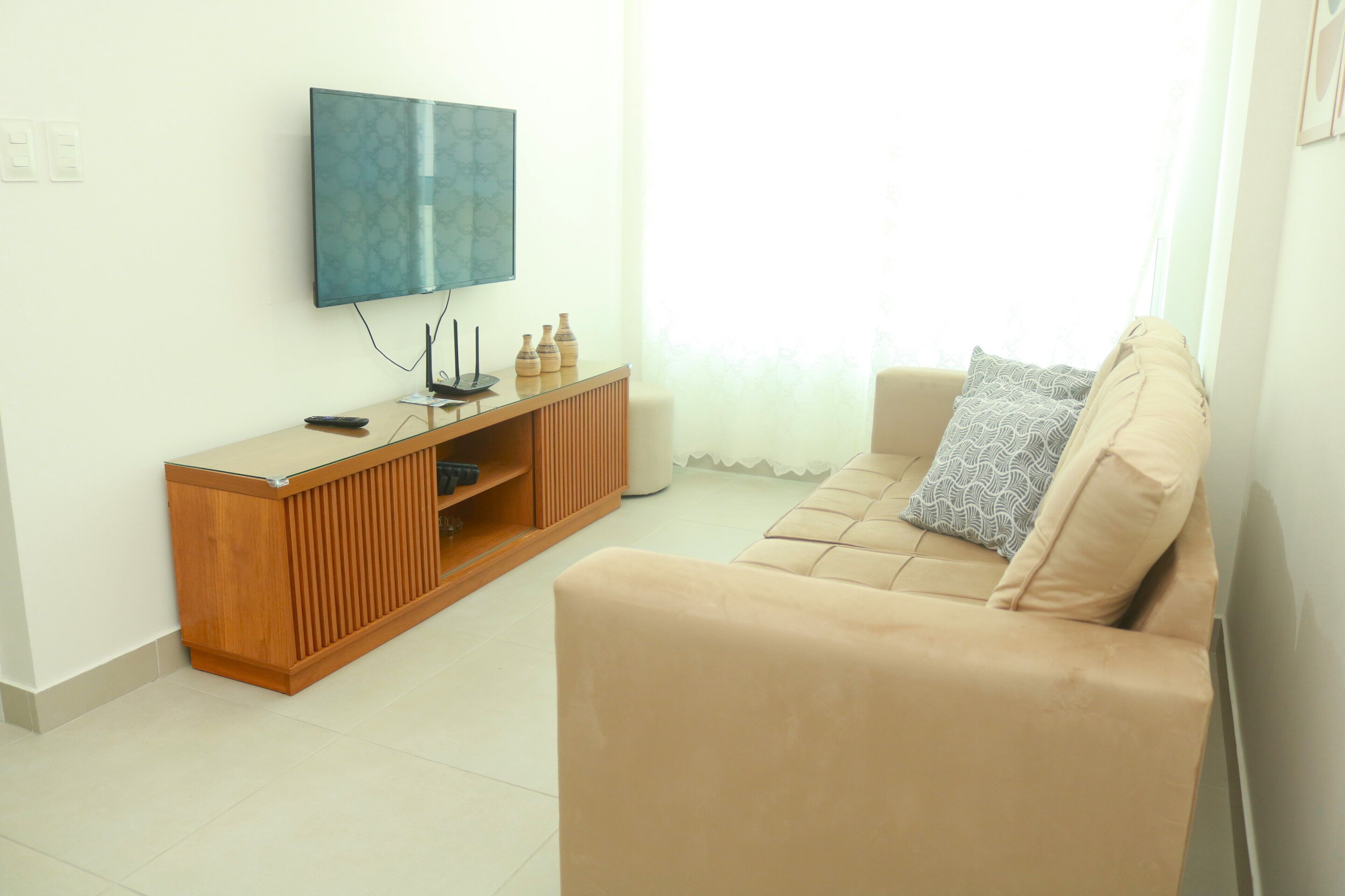 A living room with a beige sofa, a wooden entertainment unit, a flat-screen TV, and a window with sheer curtains.
