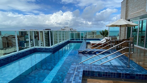 A rooftop infinity pool with a view of the ocean, lounge chairs, and a glass railing.