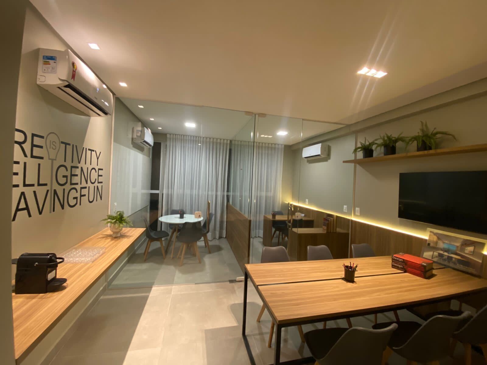 A modern office space with a glass partition, a wooden reception desk, a conference table, and a mounted TV.