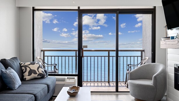 Una sala de estar moderna con un sofá gris, una mesa de centro blanca y un sillón gris. La habitación tiene un amplio ventanal corredero que da a un balcón con vistas al mar.