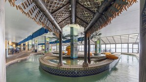 Un área de piscina interior con una fuente central, asientos alrededor de la piscina y un techo con temática tropical.
