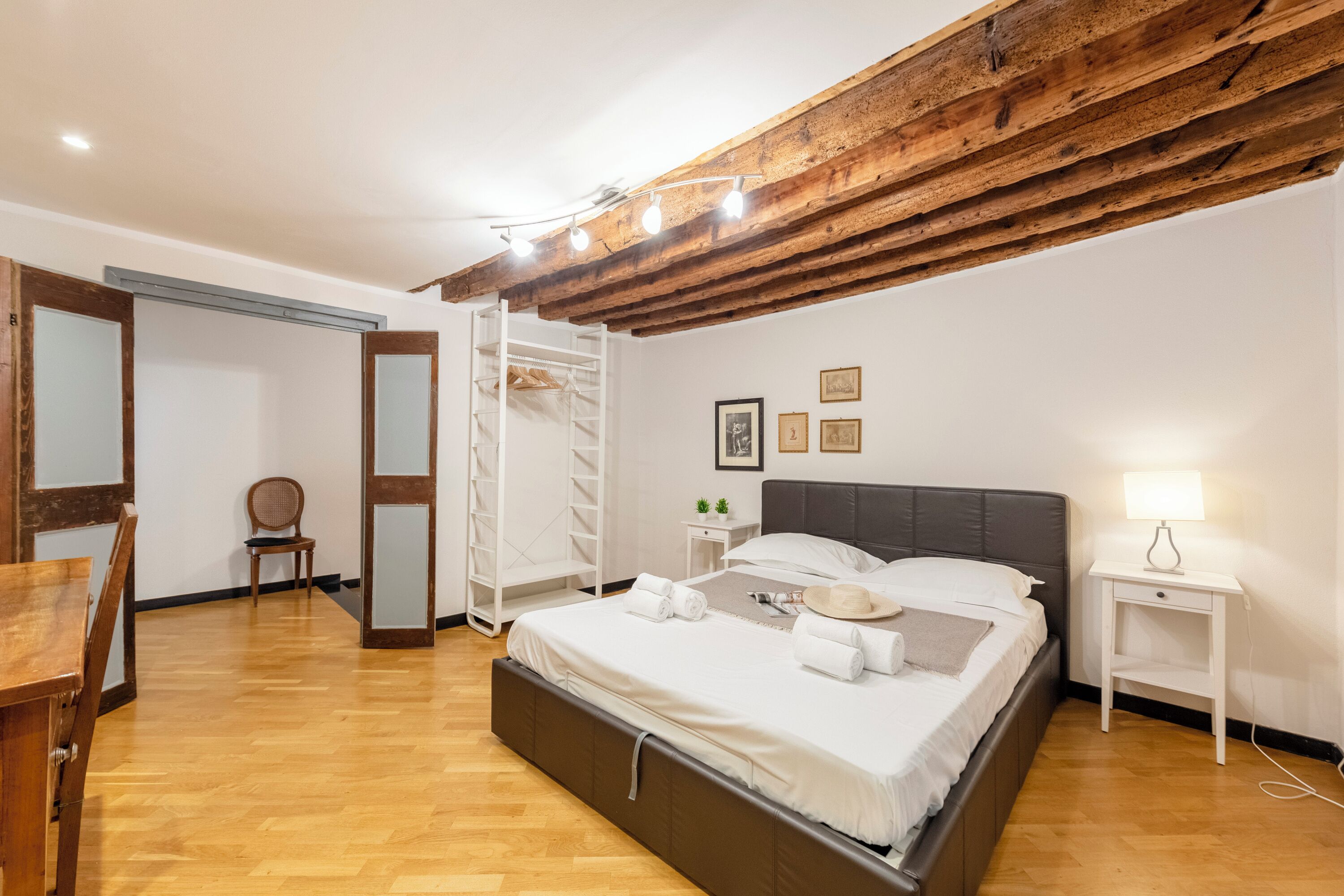 A bedroom with a wooden ceiling, a bed with white linens, a bedside table with a lamp, and a chair.
