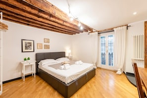 A bedroom with a wooden ceiling, a bed with white linens, a nightstand, a small table, and a window with curtains.