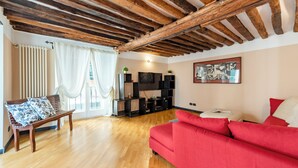 Una sala de estar con piso de madera, un sofá rojo, un banco de madera con cojines estampados, un mueble de entretenimiento negro y un techo con vigas a la vista.