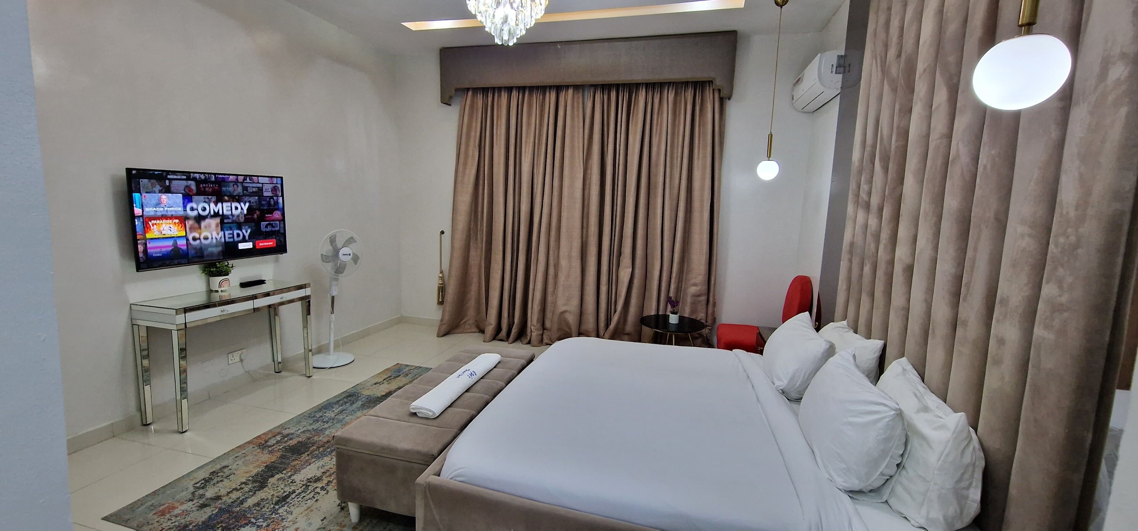 A hotel room with a bed, a TV displaying comedy channels, a desk with a fan, and a red chair.