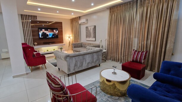 A modern living room with a grey sofa, red armchairs, a blue sofa, and a round coffee table. A flat-screen TV is mounted on the wall, and there is a floor lamp. - Luxury 3-bed House in Gated Estate With Pool Lekki (Lekki)