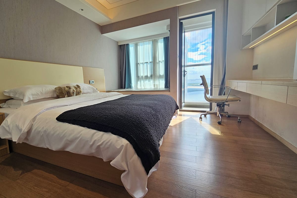Ein modernes Hotelzimmer mit einem großen Bett, einem Schreibtisch mit Lampe, einem Sessel und Blick ins Freie durch ein großes Fenster.