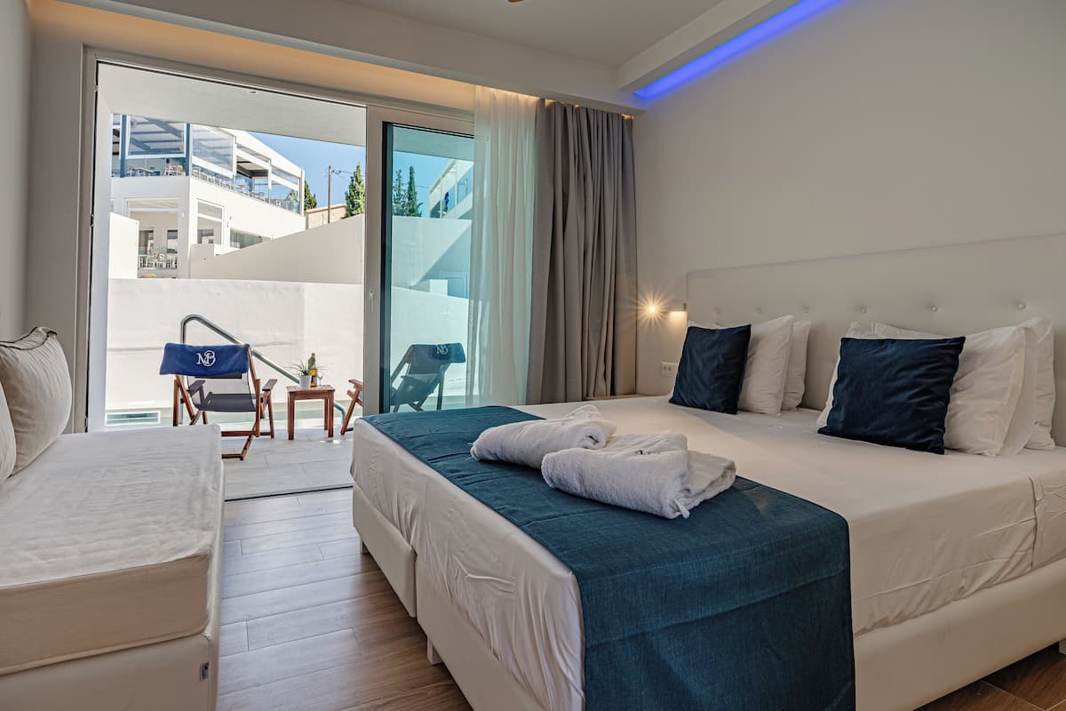 A hotel room with a bed, a sofa, a small table with a chair, and a view of a building through a glass door.