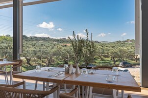 Restaurante com mesas de madeira e cadeiras de vime, com vista para oliveiras e colinas ao fundo.